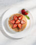 A tall stack of golden, fluffy strawberry pancakes drizzled with maple syrup on a rustic wooden table.