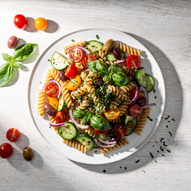 A vibrant summer pasta salad with colorful vegetables and fusilli pasta, beautifully plated on a light wood table.