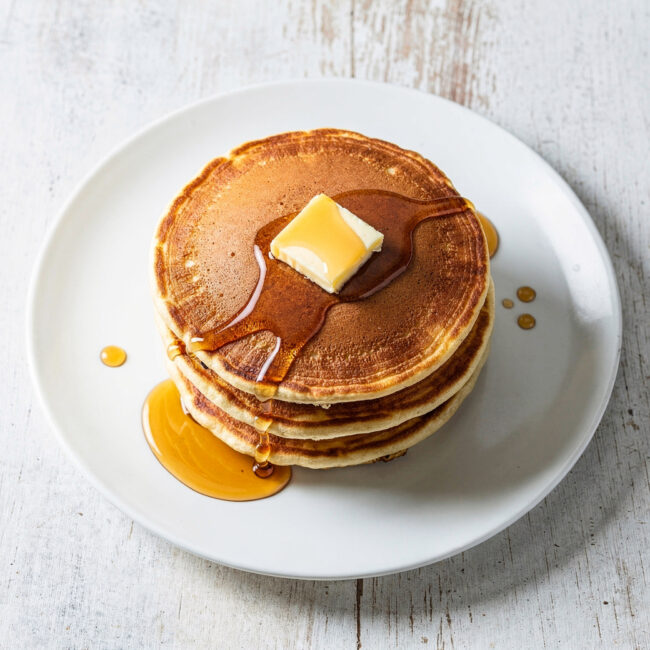 A stack of fluffy, golden pancakes from this sweet pancake recipe, drizzled with maple syrup, on a rustic wood table.