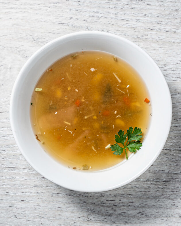 A rustic bowl of rich, golden vegetable bouillon sits on a wooden table, steam rising gently from its surface.