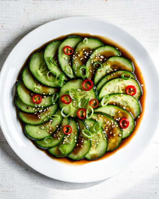 A vibrant Asian cucumber salad with thinly sliced cucumbers in a glossy dressing, garnished with sesame seeds and red chili, beautifully plated on a rustic wooden board.