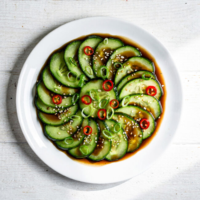 A vibrant Asian cucumber salad with thinly sliced cucumbers in a glossy dressing, garnished with sesame seeds and red chili, beautifully plated on a rustic wooden board.