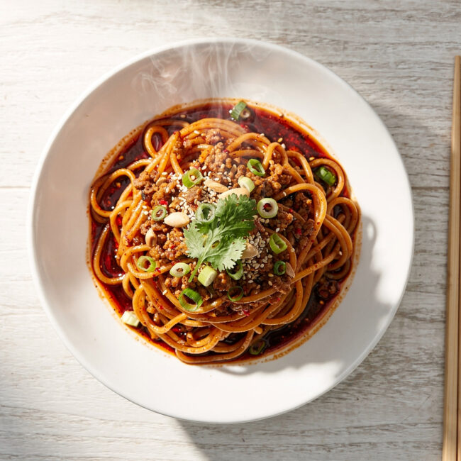 A vibrant bowl of dan dan noodles topped with ground pork, crushed peanuts, and green onions on a wooden table.