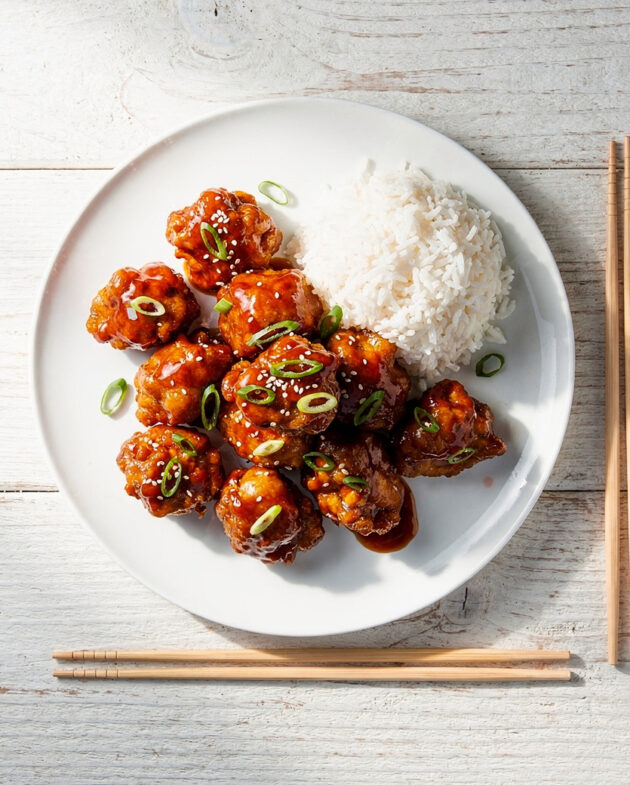 A close-up view of perfectly cooked general tso's chicken glistening with a sticky-sweet sauce on a white plate.