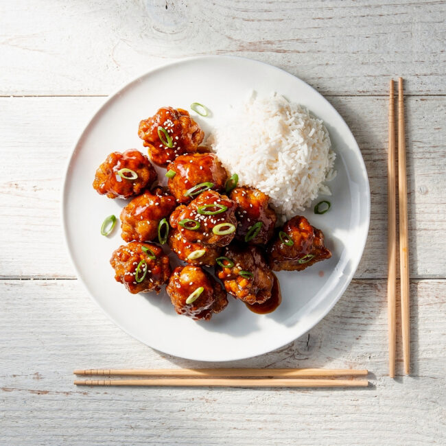 A close-up view of perfectly cooked general tso's chicken glistening with a sticky-sweet sauce on a white plate.