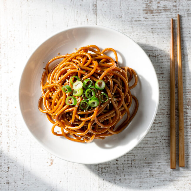 A vibrant, glossy pile of lo mein noodles tossed with colorful vegetables in a shallow white bowl.